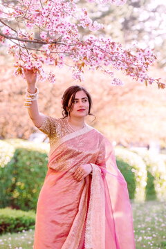 A woman in a shimmering pink traditional outfit stands serenely among cherry blossoms, radiating warmth, elegance, and cultural pride.