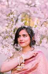 A woman in a shimmering pink traditional outfit stands serenely among cherry blossoms, radiating warmth, elegance, and cultural pride.