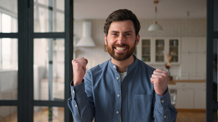 Happy joyful Caucasian guy man winner dad smiling clenching fists male winning rejoicing at home...