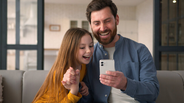 Happy positive Caucasian family daughter dad using mobile phone man guy winning child little smiling girl celebrating at home male parent typing screen victory in online game father give kid high five