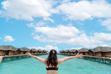 Stunning shot of a woman in a swimsuit with arms outstretched towards the beautiful ocean, framed by an idyllic overwater resort. Evokes luxury travel, relaxation, and dream vacations
