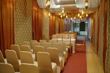 Beautifully decorated event hall with white covered chairs, a red aisle, and elegant hanging...