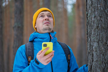 Male hiker dressed in a blue jacket and yellow beanie, looking up in a winter forest while...