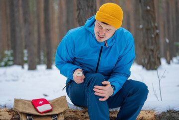 Male hiker is sitting on fallen tree trunk, holding bandage and applying it to his injured knee,...