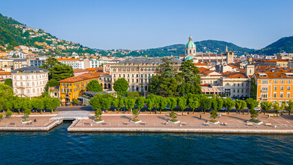 Naklejka premium Aerial view of Como village with clustered terracotta rooftops, narrow streets, lakeside promenade, and surrounding green hills under clear skies