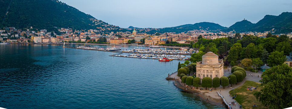 Aerial view of Volta Temple in Como, Italy, surrounded by park trees, lakefront promenade, and clear blue lake under sunny summer skies