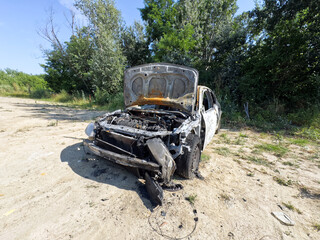 A completely burnt-out car sits on a dirt path, surrounded by green foliage under a clear sky, showing extensive damage from fire.