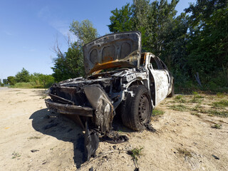A heavily burned-out car with its hood open sits abandoned on a dirt path under a clear sky,...
