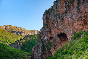 Rugged cliffside with cave. View of a rugged cliff featuring a natural cave set against lush green...