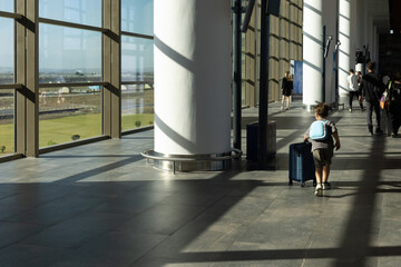 A child walks away from the camera in a sunlit airport terminal, pulling a small suitcase. The...