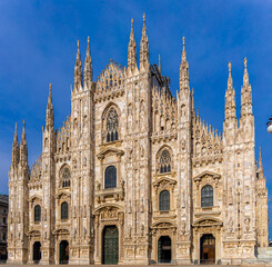 Fototapeta premium Gothic-style Milan Cathedral facade with ornate spires and intricate stonework against clear blue sky, captured in daylight