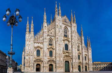 Gothic-style Milan Cathedral facade with ornate spires and intricate stonework against clear blue...