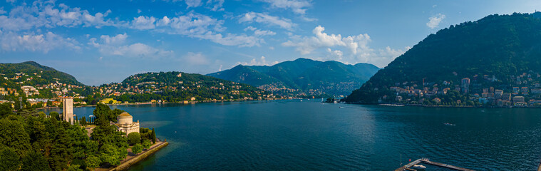 Fototapeta premium Aerial summer view of Lake Como with deep blue water, surrounding green hills, villas, and boats under a clear, sunny sky in Italy