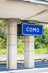 Fototapeta premium Como train station platform in Italy with overhead sign reading “Como,” railway tracks, and shelter under a bright summer sky