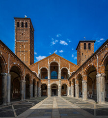 Obraz premium Courtyard view of Basilica di Sant’Ambrogio in Milan with Romanesque arches, brick facade, and twin bell towers under a vivid blue sky