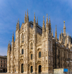 Gothic-style Milan Cathedral facade with ornate spires and intricate stonework against clear blue...