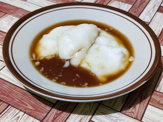 Bubur sumsum, traditional Indonesian food. Soft white marrow porridge served on a plate sprinkled with thick brown sugar sauce on a wooden patterned table.