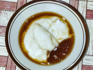 Bubur sumsum, traditional Indonesian food. Soft white marrow porridge served on a plate sprinkled with thick brown sugar sauce on a wooden patterned table.
