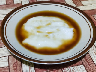 Bubur sumsum, traditional Indonesian food. Soft white marrow porridge served on a plate sprinkled with thick brown sugar sauce on a wooden patterned table.