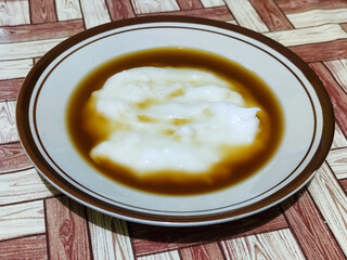 Bubur sumsum, traditional Indonesian food. Soft white marrow porridge served on a plate sprinkled with thick brown sugar sauce on a wooden patterned table.