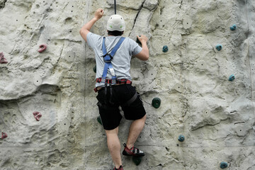 An individual ascends an artificial rock climbing wall wearing safety equipment and a harness