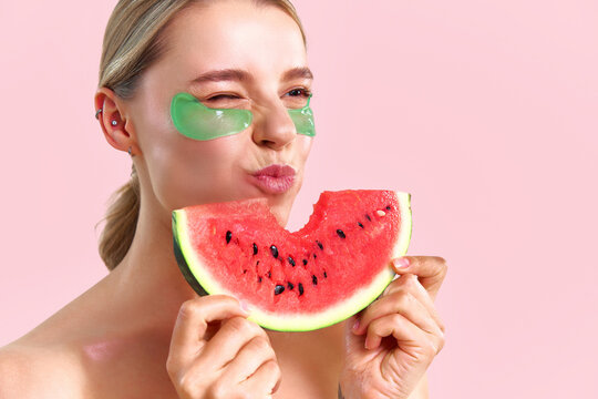 A delicious, juicy watermelon. A beautiful girl wearing green under-eye patches is eating watermelon against a soft pink background. - Powered by Adobe