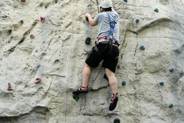 An individual ascends an artificial rock climbing wall wearing safety equipment and a harness
