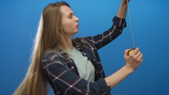 Woman measuring with tape against isolated blue wall showing precision and focus in casual attire, reflecting practicality and concentration in a simple studio setting.