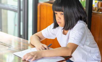 Asian girl with black hair sitting at table thinking deeply while studying. Wearing white shirt,...