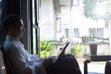 Asian businessman sitting by window relaxing with iced coffee and using digital tablet in cafe. Morning sunlight, remote working lifestyle, online reading, focus, calm, and leisure concept