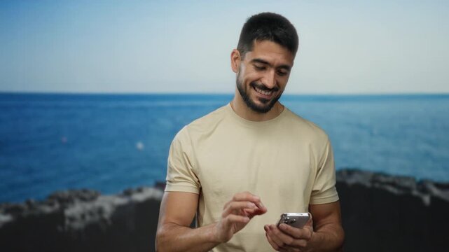 Young man smiling at smartphone by the seaside, capturing a relaxed moment at the beach with the scenic ocean backdrop enhancing his stylish and casual vibe.