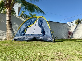 Tenda de campismo montada num relvado tropical sob uma palmeira num dia ensolarado. Ambiente tranquilo e perfeito para lazer ao ar livre.