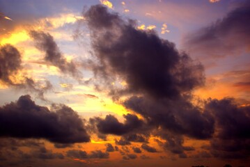 An intense sky at dawn or dusk where dark, billowing clouds are pierced by vibrant orange and...