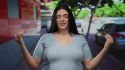 Woman meditating outdoors in a city street poses peacefully wearing casual attire with hands together, embracing tranquility surrounded by urban trees, cars, and buildings.