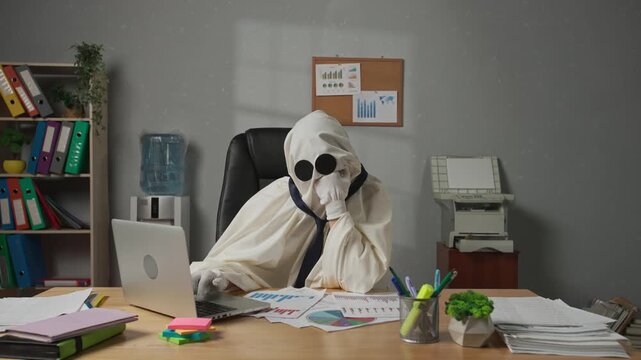 Halloween businessman ghost in white sheet sits at office desk works on laptop, tired bored expression, sighing and thinking. Happy halloween business concept.