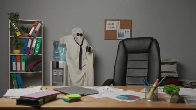 Halloween businessman ghost in white sheet standing in office drinking water, taking break during busy day. Happy halloween business concept.