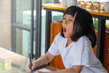 Asian young girl yawns widely holding a pen, clearly tired during a study session. Sitting at a wooden table , capturing a candid moment of learning fatigue in a cozy home