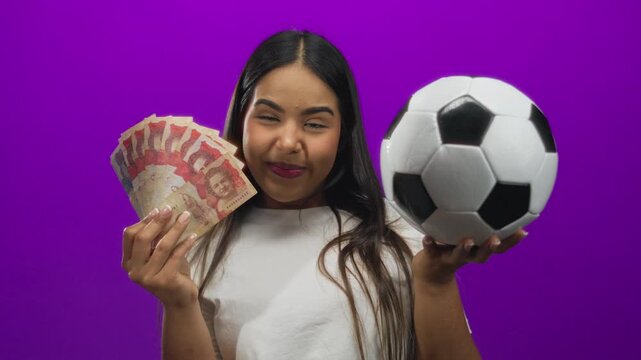 Woman smiling holding colombian pesos and soccer ball against vibrant purple background representing latin culture and sport enthusiasm.