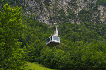 A modern cable car glides above a vibrant green forest, with a steep, rocky mountain face rising in...