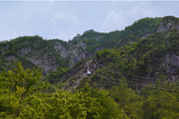 A lone cable car ascends a lush green mountain, offering scenic views of the surrounding peaks and valleys under a soft, cloudy sky.