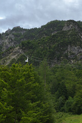 A solitary cable car ascends a lush green, forested mountainside under a cloudy sky, with buildings perched near the summit.