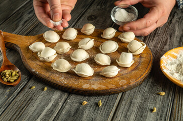 A person sprinkles flour over neatly arranged dumplings on a wooden board. The kitchen is warmly...
