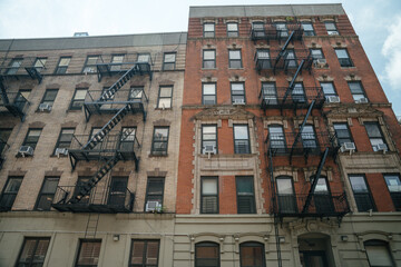 Two classic brick apartment buildings stand side by side, each featuring black metal fire escapes. Window air conditioners and architectural details emphasize the New York City residential style.