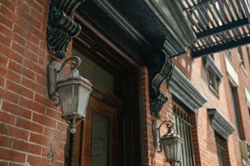 Two classic wall lanterns are mounted on a red brick facade, framed by black carved corbels and an ornate entrance. A metal fire escape casts shadows along the traditional architecture.