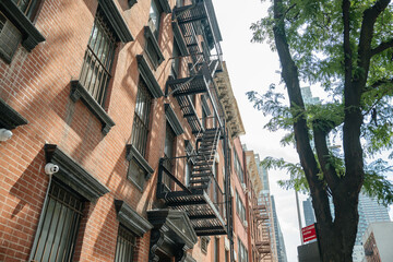 Black metal fire escapes line a red brick apartment building under bright sunlight. A large tree and city skyscrapers add contrast to the traditional urban architecture.