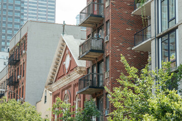 A red-brick church roof with classical trim stands beside old and new apartment buildings. Balconies, fire escapes, and high-rises contrast in this layered urban cityscape.