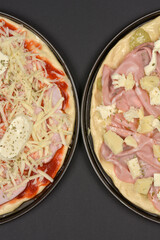 Two unbaked pizzas, one with ham and cheese and the other with mortadella and cheese, are positioned side-by-side on a dark surface, ready for baking