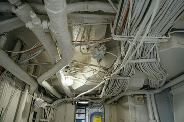 A close-up view of the ceiling in a military ship shows a dense network of pipes and bundled wires....