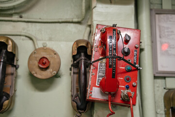 Close-up of a red military communication phone with labeled buttons and a corded handset. The panel...
