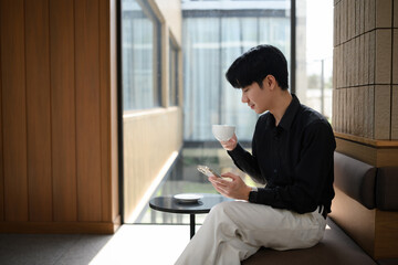 An Asian man has a casual coffee break while browsing on his mobile in a light-filled modern space